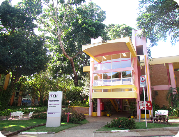 Predio rosa e amarelo com uma placa ao lado esquerdo escrito IFCH, muitas árvores ao redor.