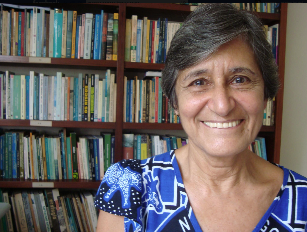 Mulher sorrindo, com cabelos lisos e curtos e roupa azul de mangas curtas, com estante de livros ao fundo. 