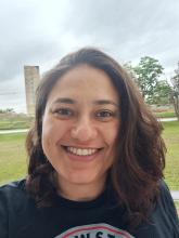 Mulher sorrindo, com cabelos castanhos e ondulados na altura dos ombros, vestindo camiseta preta. Ao fundo um gramado, um prédio, poucas árvores e céu nublado.